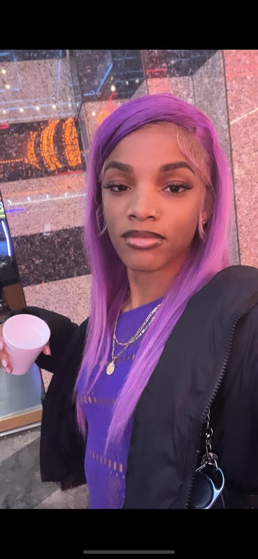 Young woman with long purple hair posing confidently indoors, holding a cup and wearing a dark jacket.