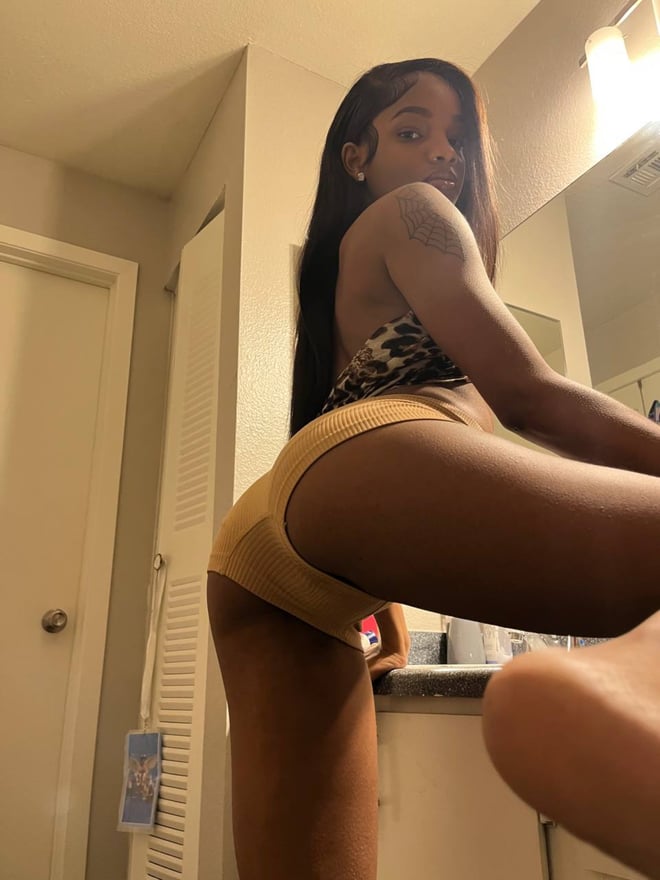 Young woman with long dark hair posing in front of a mirror in a bathroom, wearing a leopard-print top and yellow shorts while showing her figure.