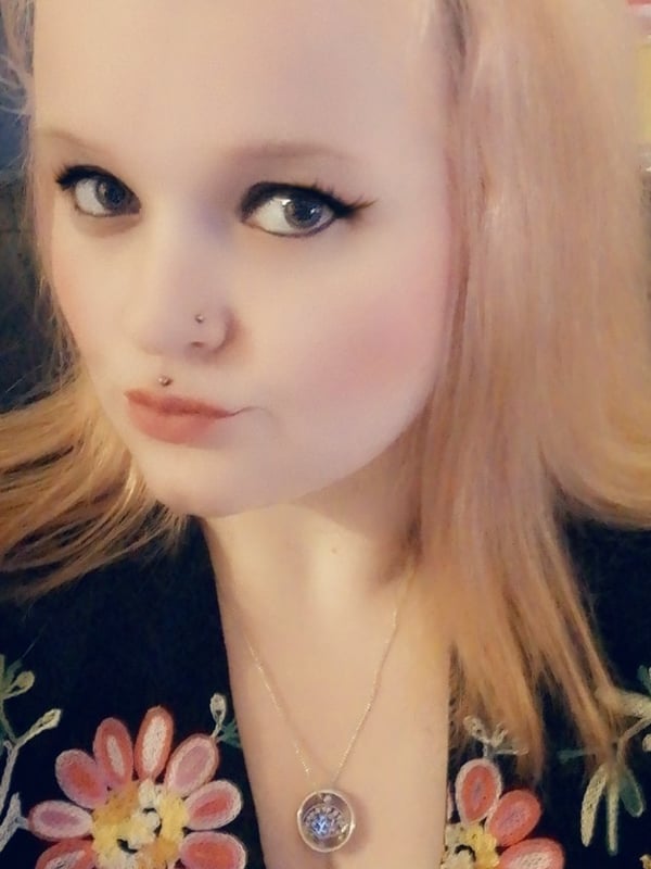 A woman with long blonde hair wearing a black floral top and a necklace, posing with a close-up facial expression.