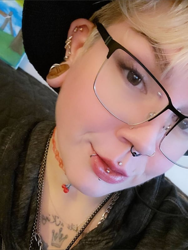 Close-up portrait of a person with short blonde hair, glasses, facial piercings, and earrings, wearing a hat and necklace.