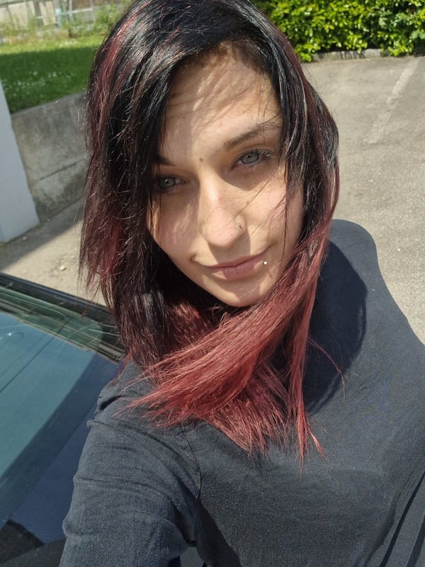 Young woman with dark red hair taking a selfie outdoors near a building and greenery.