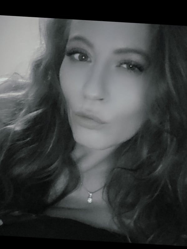 Black-and-white close-up selfie of a woman with wavy hair, subtle makeup, and a necklace, gazing at the camera.