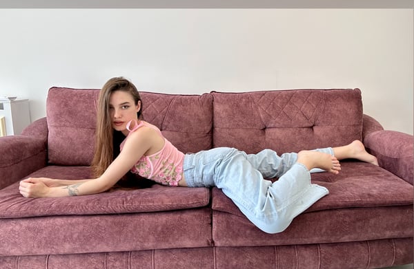 Young woman with long brown hair lounging on a pink couch, wearing a pink top and light blue pants, posing relaxedly with bare feet.