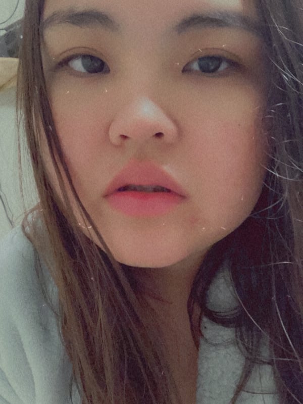 A young woman with long dark hair taking a close-up selfie, smiling softly while wearing a light-colored top.