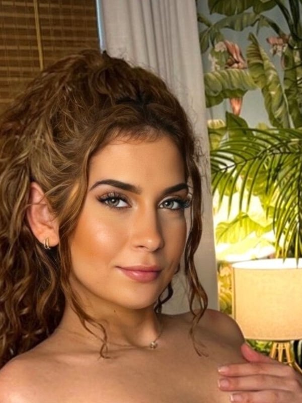 Young woman with curly brown hair smiling at the camera in a tropical indoor setting with plants and warm lighting.