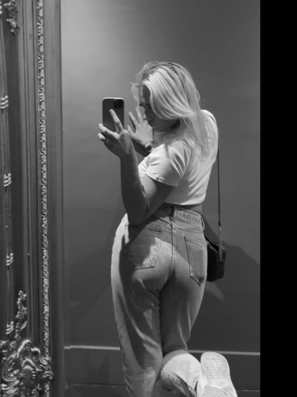 Blonde woman taking a mirror selfie in a white shirt and light pants in front of an ornate mirror.