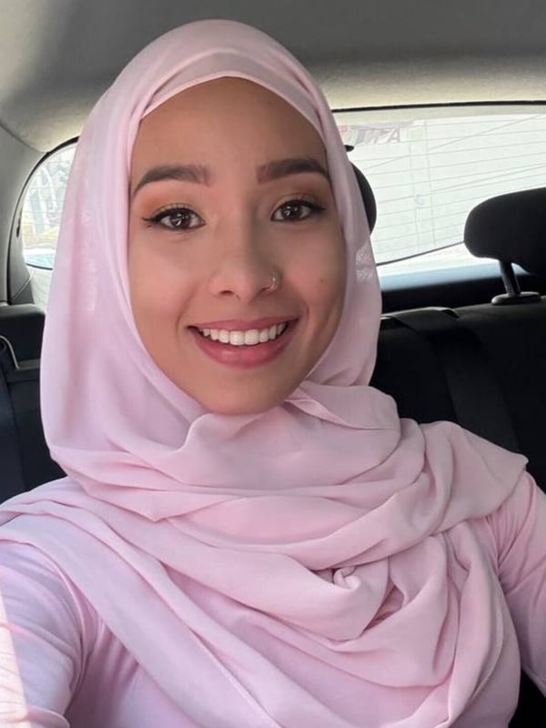 A smiling young woman wearing a pink headscarf and top, sitting in a car with a window in the background.