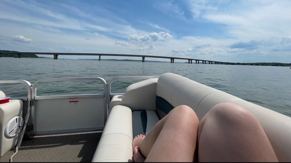 A woman relaxing on a boat with legs extended, overlooking a lake and distant bridges under a clear sky.