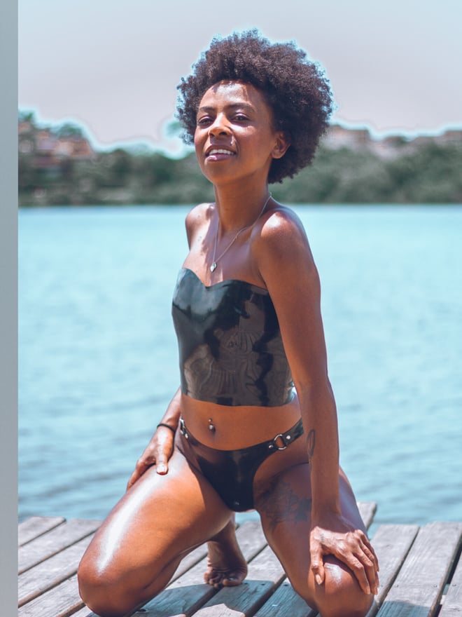 A woman smiling while kneeling on a wooden dock by the water, dressed in a strapless top and bottoms with a belly piercing.