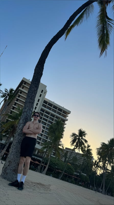 Young man standing shirtless by a palm tree on a beach at sunset, with a hotel building and ocean in the background.