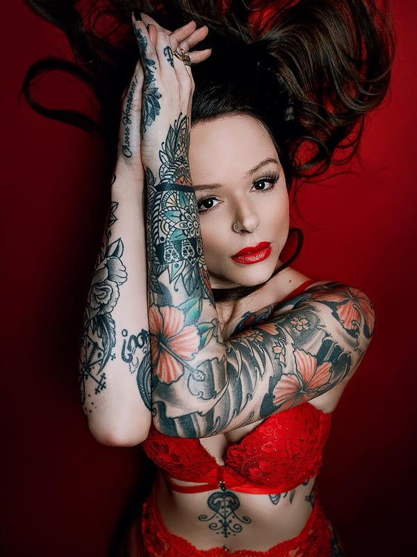 A tattooed woman with flowing dark hair lies back against a red background, wearing red lingerie and displaying intricate arm tattoos.