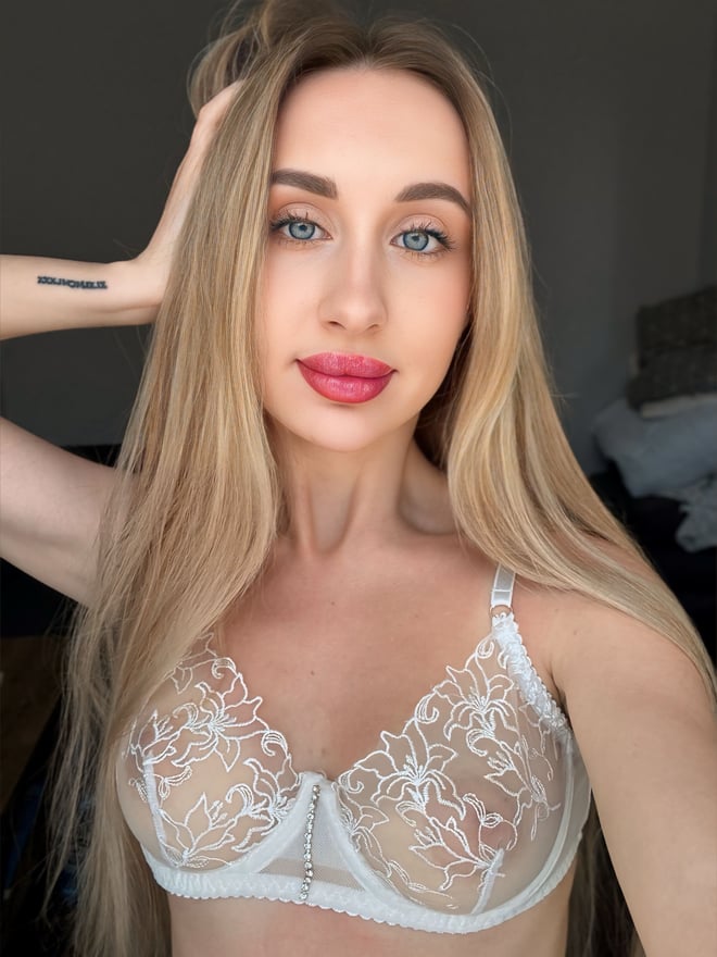 Close-up of a woman with long blonde hair and blue eyes, hand on her head, wearing a white lace bra, smiling with red lipstick.