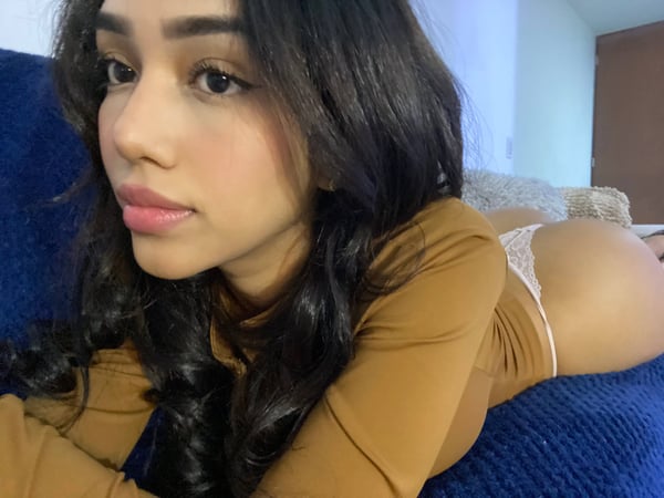 Young woman with long dark hair lying on a blue couch in a bedroom, wearing a long-sleeve top and underwear, posing sideways with a soft smile.