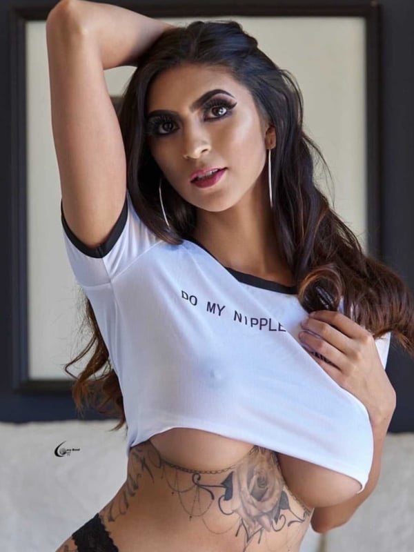 A woman with long dark hair posing confidently in a cropped white t-shirt and black bottoms, standing against a framed background.