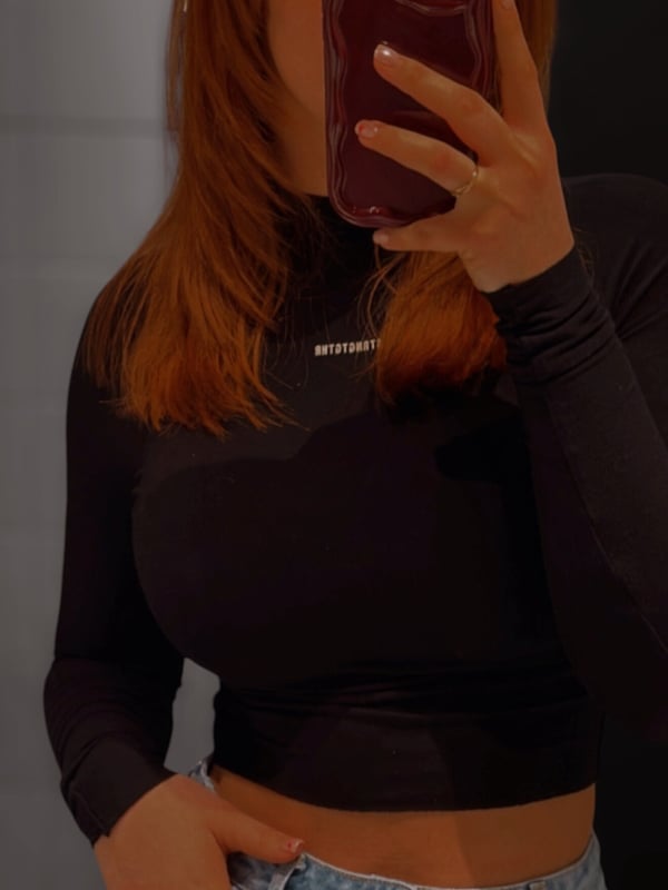 A woman taking a mirror selfie in a black turtleneck top and light blue jeans, holding her red iPhone with reddish-brown hair.