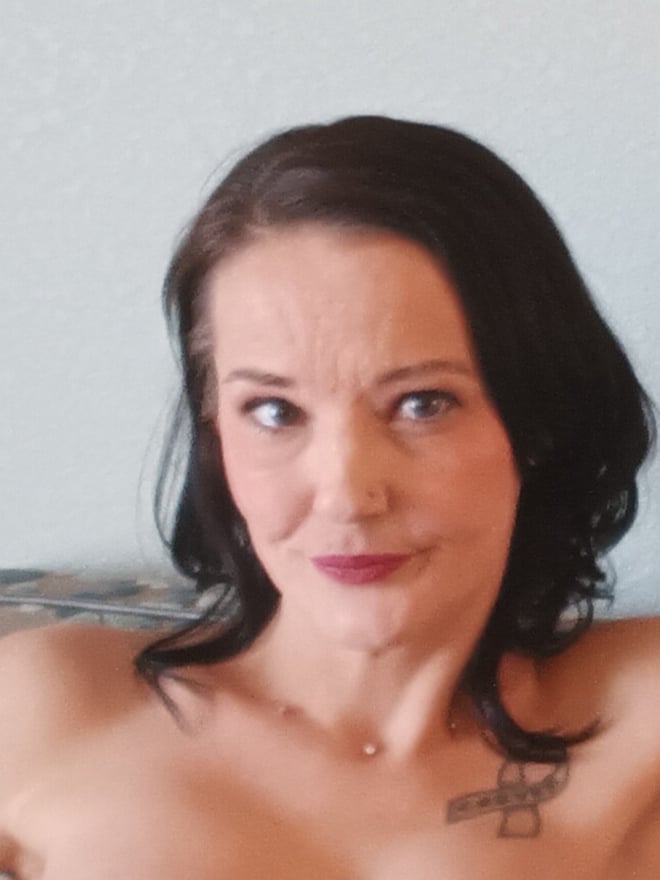 A woman with dark wavy hair and a necklace poses shirtless on a gray couch, smiling at the camera.