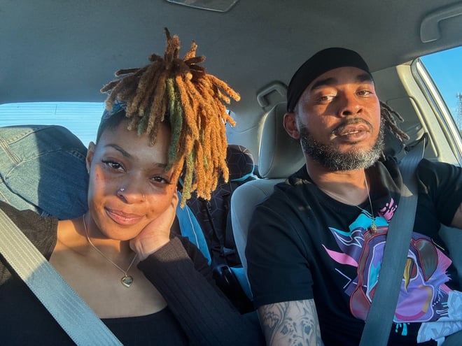 A young woman with colorful dreadlocks and a man wearing a cap pose closely together inside a car, smiling at the camera with a scenic outdoor view through the windows.