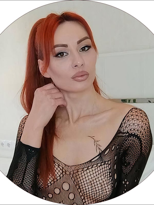 A woman with long red hair in a ponytail, wearing sheer black lingerie, posing confidently indoors.