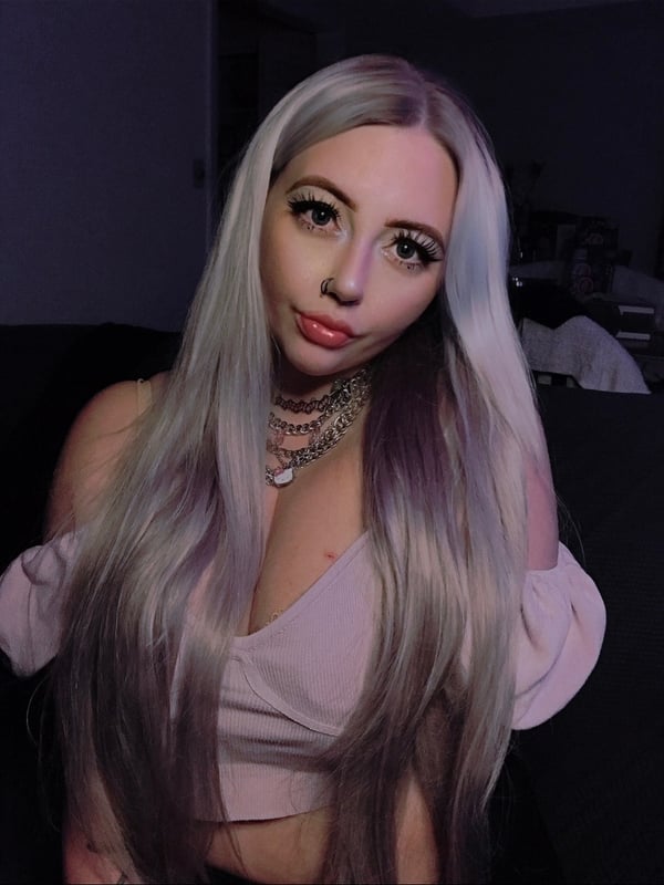 A young woman with long platinum hair wearing a pink off-shoulder top and choker necklace, posing with a sultry expression in low light.