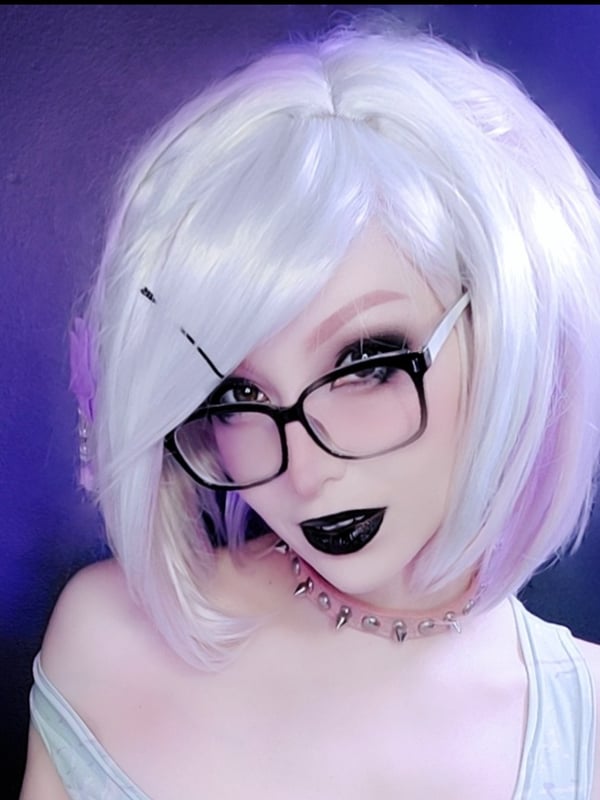 Close-up portrait of a person with white bob hairstyle, glasses, and dark lipstick, wearing a choker against a purple background.
