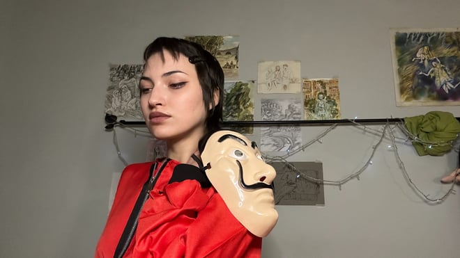 A young woman with short dark hair wearing a red jumpsuit poses with a white mask held to her shoulder in an artistic room with wall art and hanging decor.