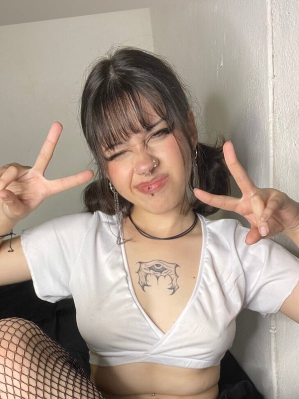 Young woman with bangs and pigtails making a playful hand gesture, wearing a white top and fishnets, smiling against a plain wall.
