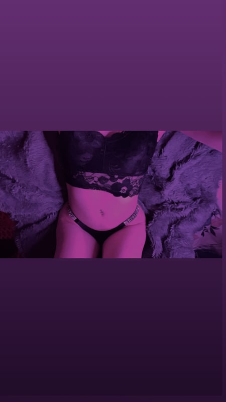 A woman posing in black lace lingerie with purple lighting and fur accents in the background.