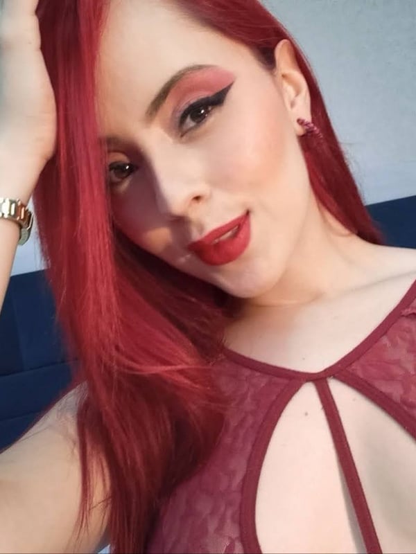 A woman with long red hair and bold makeup posing confidently, wearing a lace top and gold watch in an indoor setting.