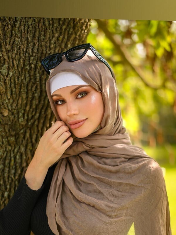 A woman wearing a headscarf and sunglasses poses thoughtfully against a tree in a verdant outdoor environment.
