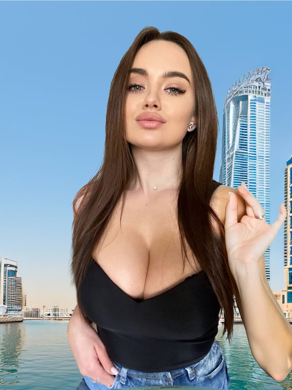 A woman with long dark hair smiling at the camera, wearing a black top, against a skyline of modern skyscrapers and water in Dubai.