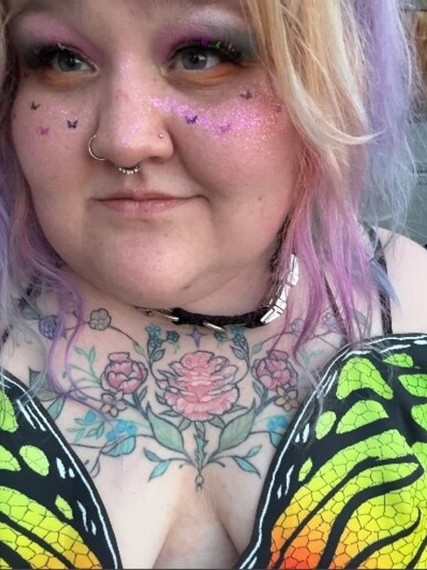 Close-up portrait of a woman with vibrant purple hair, facial piercings, and tattoos, wearing a colorful patterned top and choker necklace outdoors.