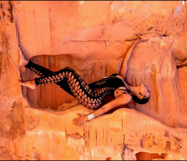 A woman posing dynamically in a black mesh outfit inside a rocky cave with warm lighting.