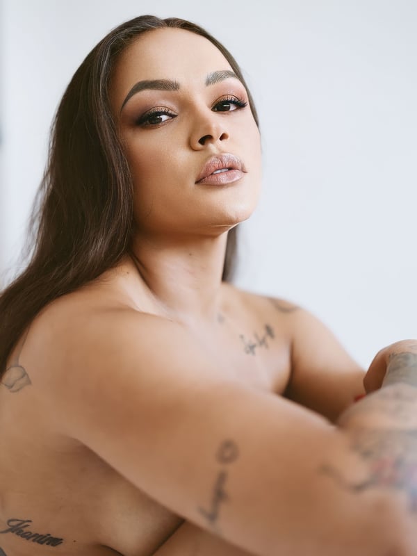 Close-up portrait of a woman with long dark hair, striking makeup, and visible tattoos on her arms.
