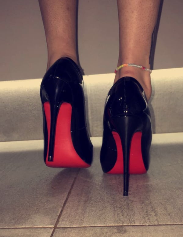Close-up view of a person's legs wearing black high-heeled shoes with red soles, standing on a tiled floor.