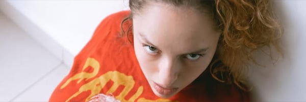 A young woman with curly red hair wearing an orange shirt, looking up at the camera with a playful expression.