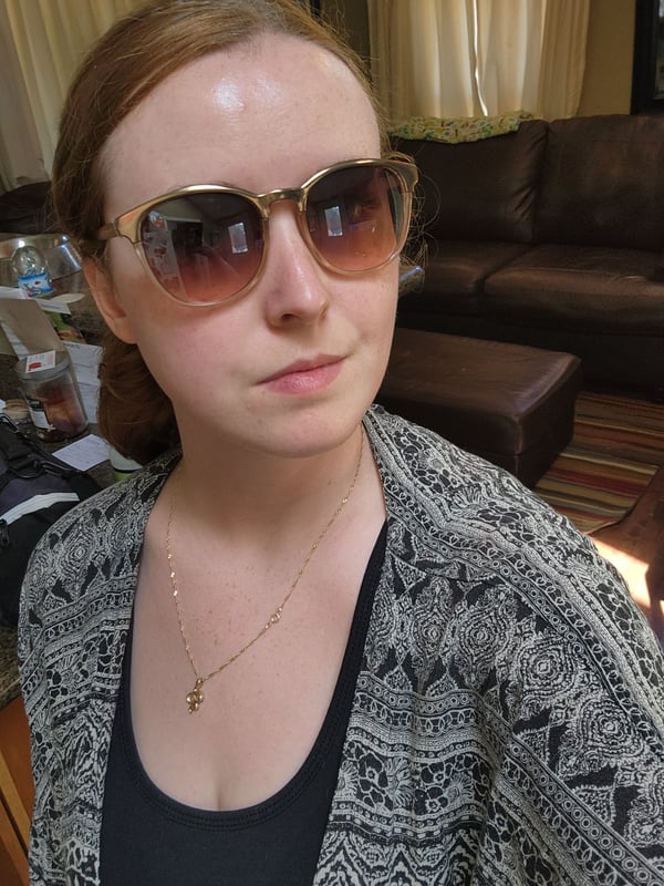 Young woman with reddish hair in a ponytail, wearing large gradient sunglasses and a patterned cardigan, posing indoors with a neutral background.