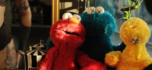 A person with tattoos posing playfully with Elmo, Cookie Monster, and Big Bird Sesame Street puppets in a stylish indoor setting with plants and a mirror.