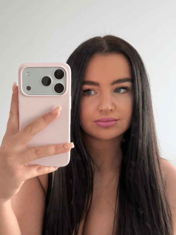 A close-up selfie of a woman with long dark hair holding a pink-cased phone in front of her face.