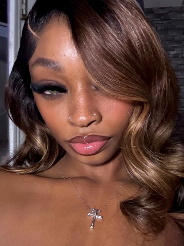 Close-up portrait of a young woman with long wavy brown hair, bold eye makeup, glossy lips, and a cross necklace, posing indoors near a door.