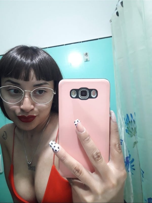 A woman with dark bangs and glasses takes a close-up selfie in a bathroom, holding a pink phone case while wearing a red swimsuit top.