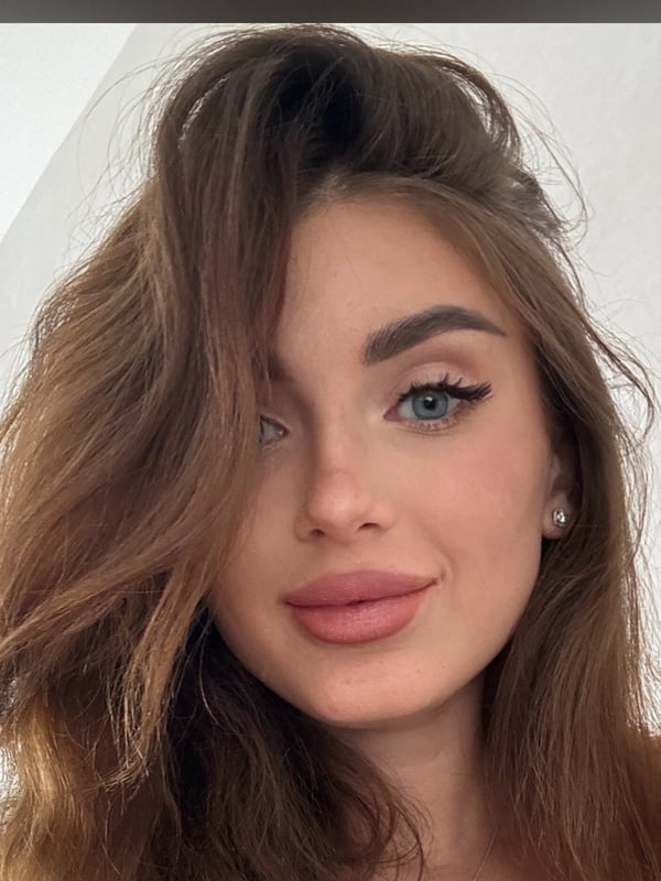 Close-up portrait of a young woman with wavy brown hair, blue eyes, and makeup, smiling softly indoors.