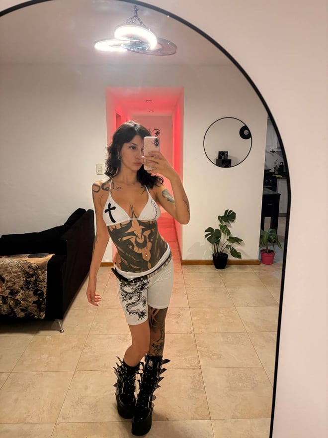 A tattooed woman with dark hair poses in black lingerie and gloves while looking toward the camera.