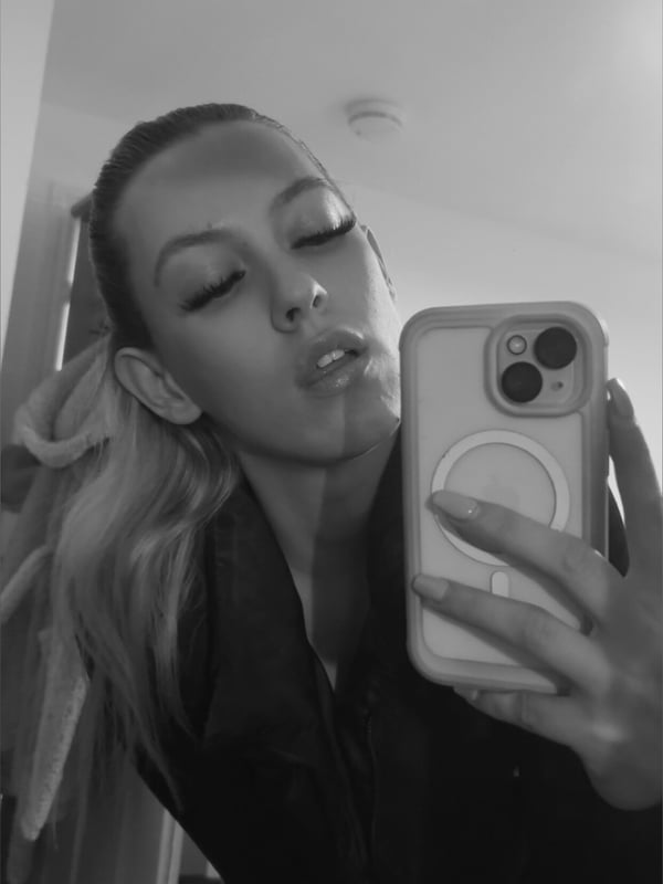 Young woman with long blonde hair making a playful expression, holding her phone for a black-and-white selfie indoors.