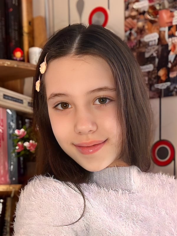 Close-up of a young woman with straight dark hair, hair clips, and a gentle smile, wearing a soft sweater in a warmly decorated indoor setting.