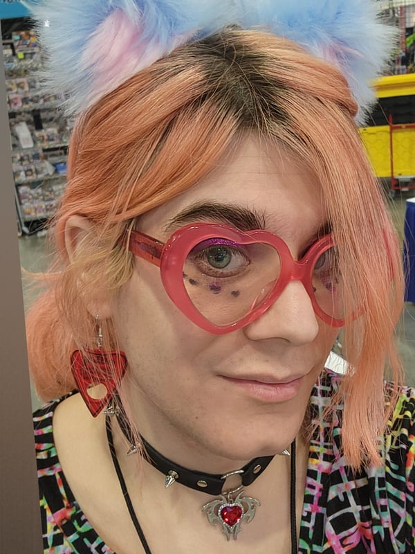 A person with dyed pink hair and cat ear headband, wearing heart-shaped glasses and a choker, posing in a store aisle.