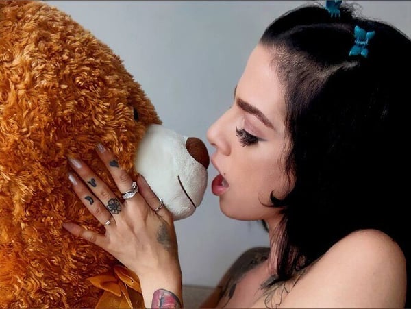 A woman with dark hair and blue hair clips kisses a large teddy bear while holding it close.
