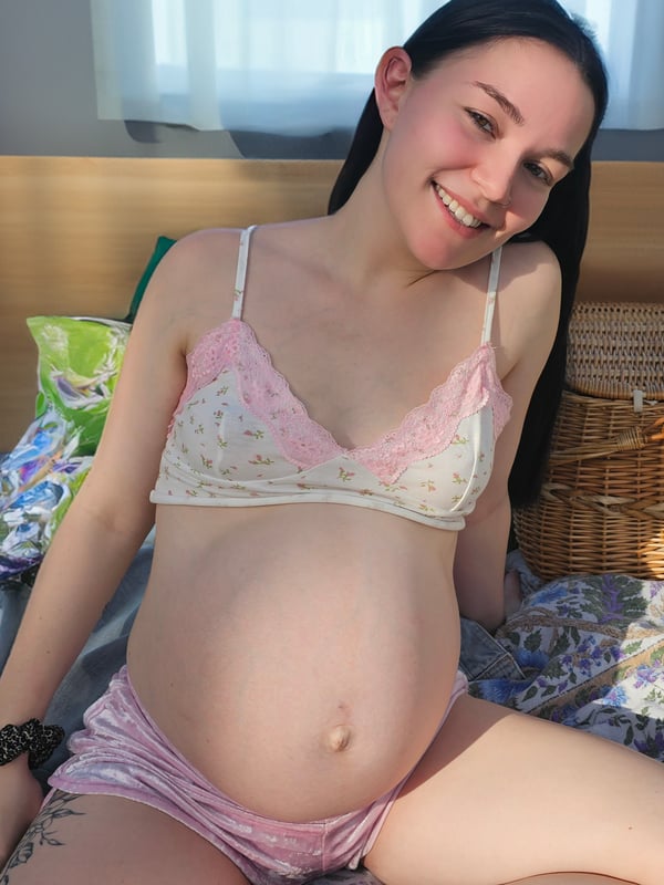 A pregnant woman sitting on a bed, smiling warmly at the camera while wearing lace lingerie.