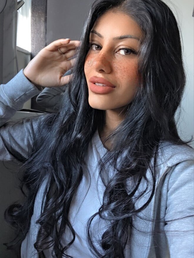 A young woman with long dark hair smiling at the camera, wearing a light blue hoodie, standing in front of a mirror in a bathroom.