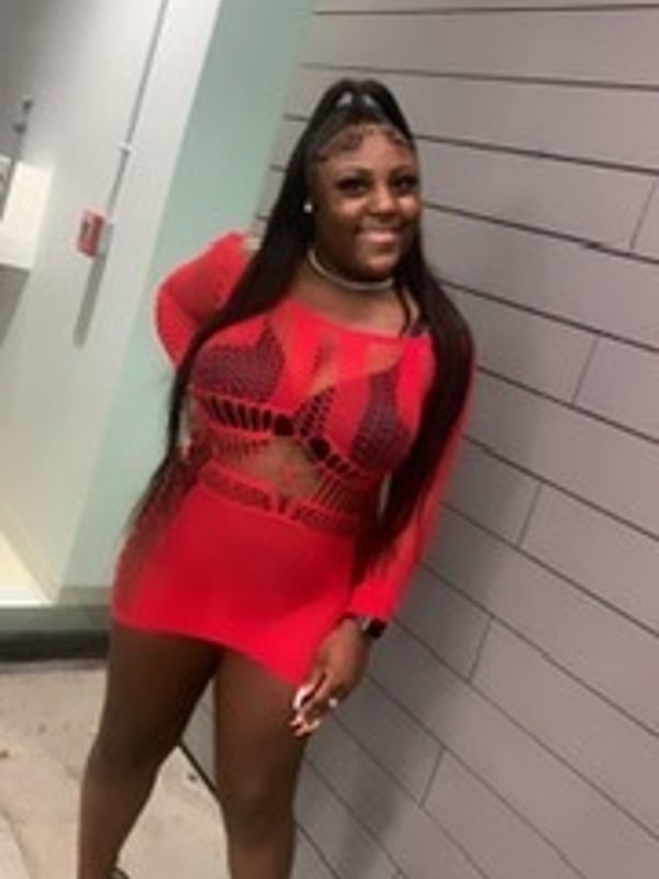 Young woman with long braided hair smiling in a form-fitting red dress against a tiled bathroom wall.