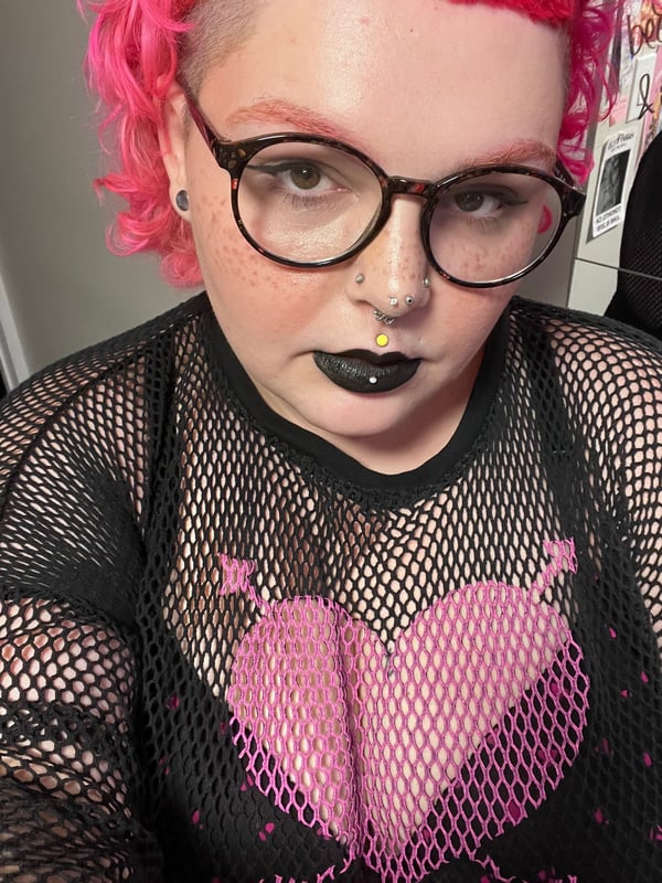 Woman with pink hair and glasses wearing a black mesh top, posing indoors with a serious expression.